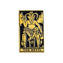Gold-tone The Devil tarot pin with a horned central figure, smaller figures below and title lettering inside a rectangular frame.