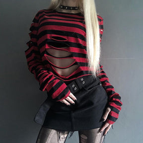 Red and black ripped crop sweater photographed with one hand against the skirt, showing the staggered front cutouts beneath the round neckline.