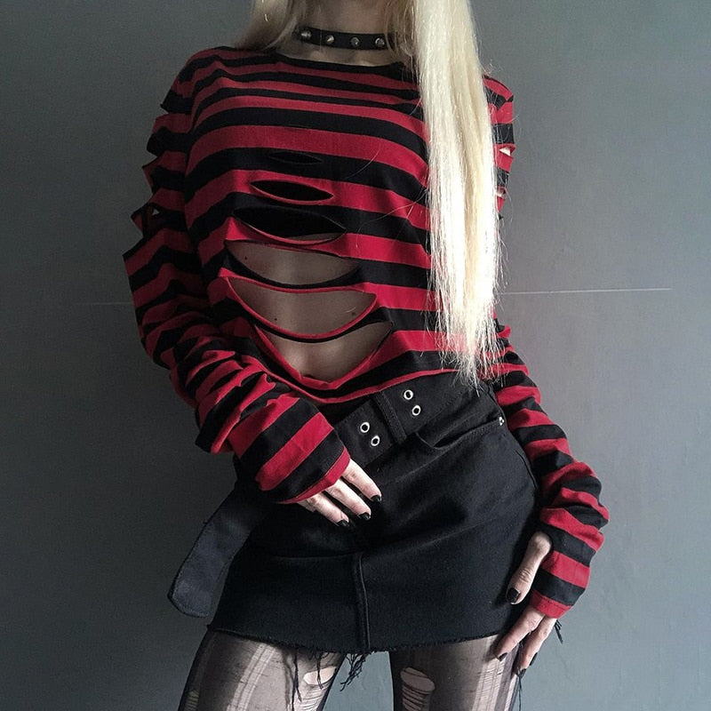 Red and black ripped crop sweater photographed with one hand against the skirt, showing the staggered front cutouts beneath the round neckline.