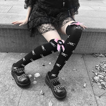 Black thigh-high stockings with scattered white skulls and pink bows, shown seated on a ledge.
