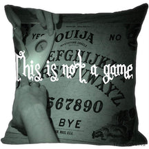 Gray square cushion cover showing hands around a printed Ouija board beneath the white phrase This is not a game.
