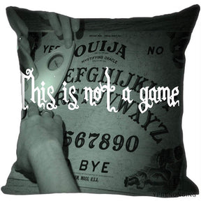 Gray square cushion cover showing hands around a printed Ouija board beneath the white phrase This is not a game.