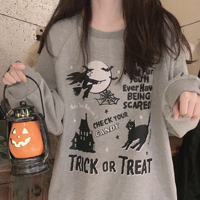 Close front view of the gray sweatshirt's flying witch, moon, spiderweb, haunted castle and black cat graphic beside a pumpkin lantern.