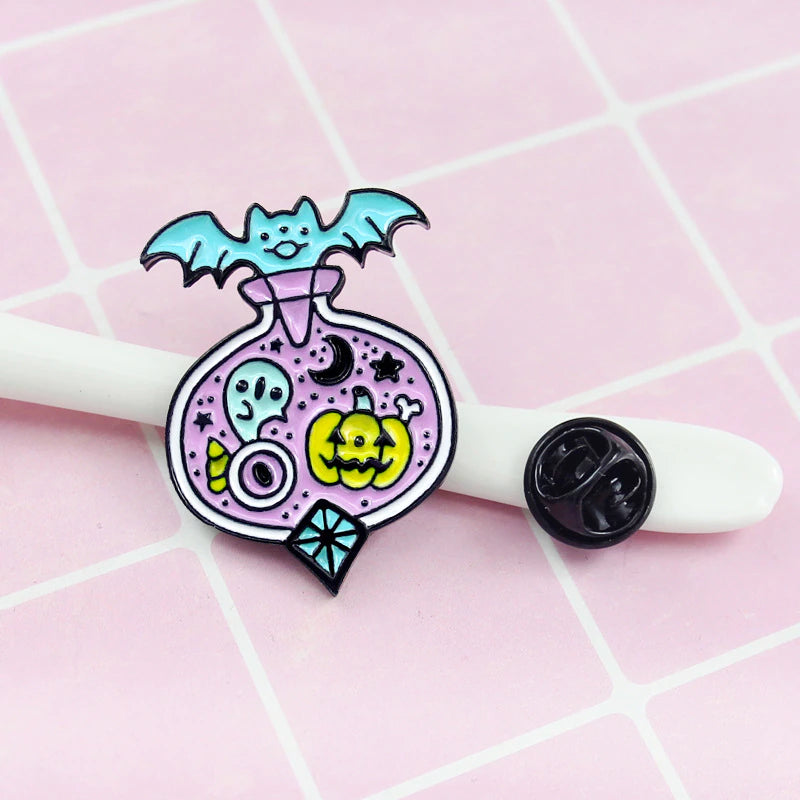 Halloween potion-bottle enamel pin beside its black backing on a white prop against a pink grid. The front shows a turquoise bat and ghost, pumpkin, moon and candy details.