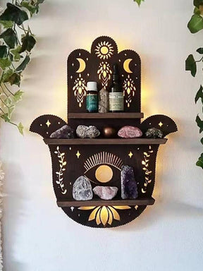 Dark brown Hamsa-shaped wooden wall shelf styled with crystals and small bottles, surrounded by trailing greenery. Sun, crescent moon, eye and lotus details decorate the back panel, with warm light glowing behind it.