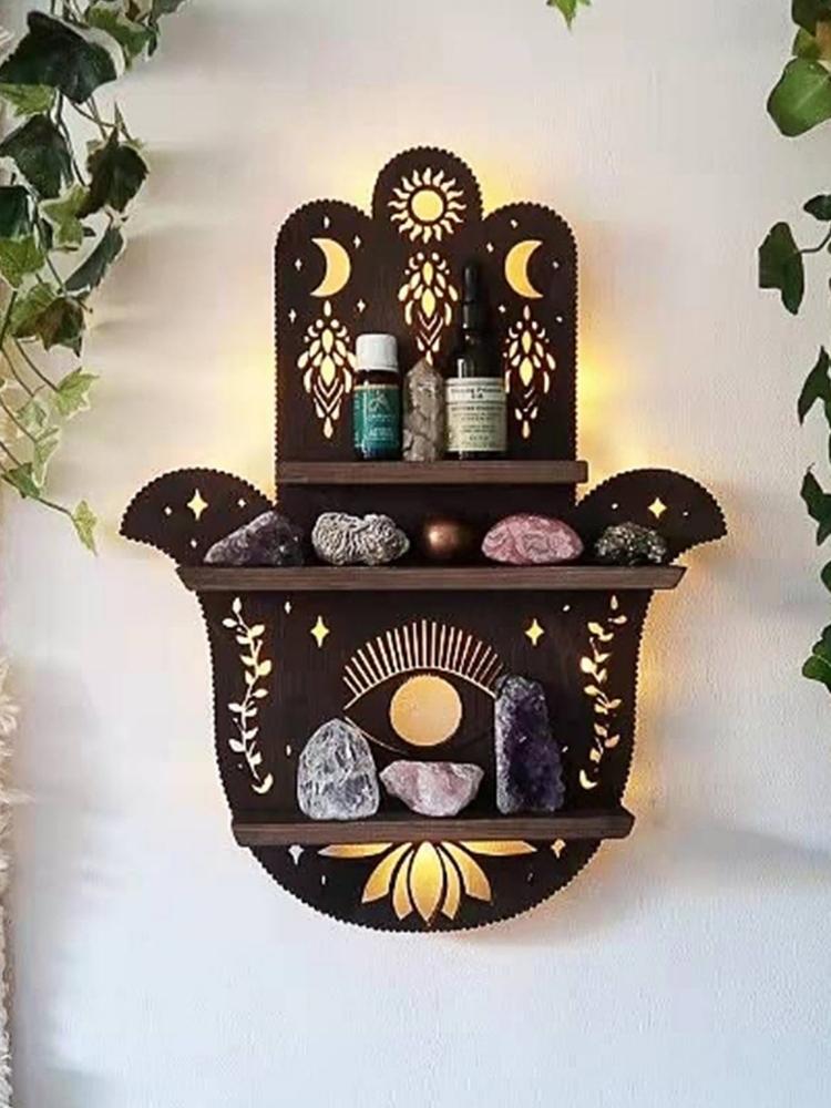 Dark brown Hamsa-shaped wooden wall shelf styled with crystals and small bottles, surrounded by trailing greenery. Sun, crescent moon, eye and lotus details decorate the back panel, with warm light glowing behind it.