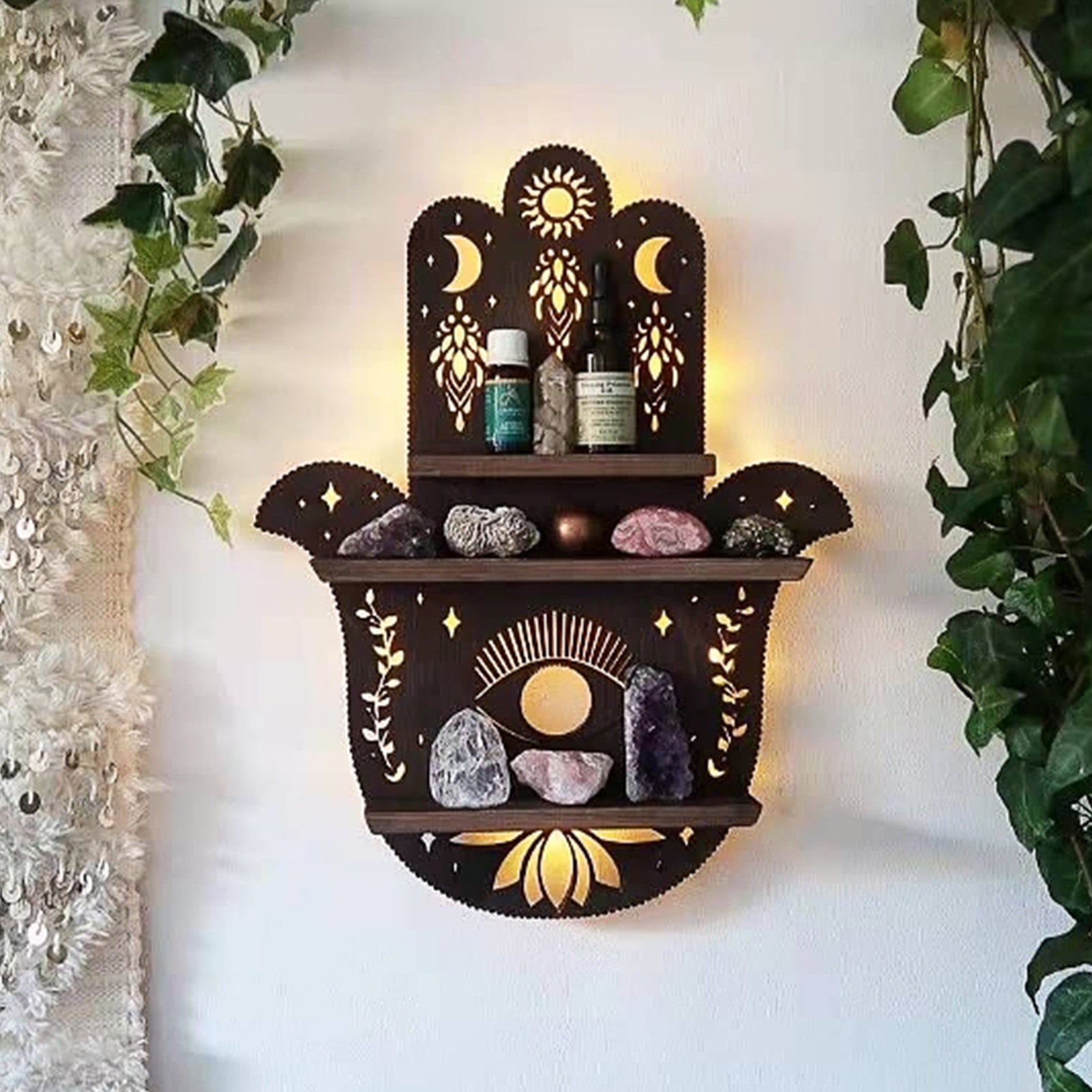 Hamsa wooden shelf mounted on a white wall between trailing green leaves, with bottles and stones arranged over the hand, eye and lotus design.