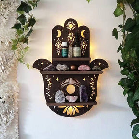 Hamsa wooden shelf mounted on a white wall between trailing green leaves, with bottles and stones arranged over the hand, eye and lotus design.