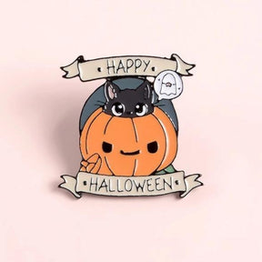 Happy Halloween cat-and-pumpkin enamel pin photographed on a pale pink background, with a white ghost beside the upper banner.
