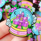 Pastel snowglobe enamel pin held between fingers, showing a pink haunted castle, white ghosts, turquoise sky and floral base, with more pins behind it.