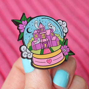 Haunted castle snowglobe pin held against a pink background, showing the crescent moon, white ghosts, green vines and pink heart on the base.