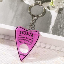 Bright pink glitter planchette keychain with black spirit-board lettering and a black border, resting on lace below its silver-tone keyring.