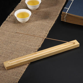 A slender incense stick rests in a long wooden holder beside teacups and stacked books on a woven mat.