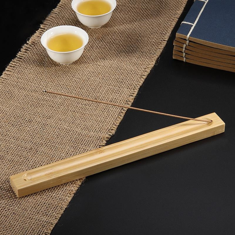 A slender incense stick rests in a long wooden holder beside teacups and stacked books on a woven mat.