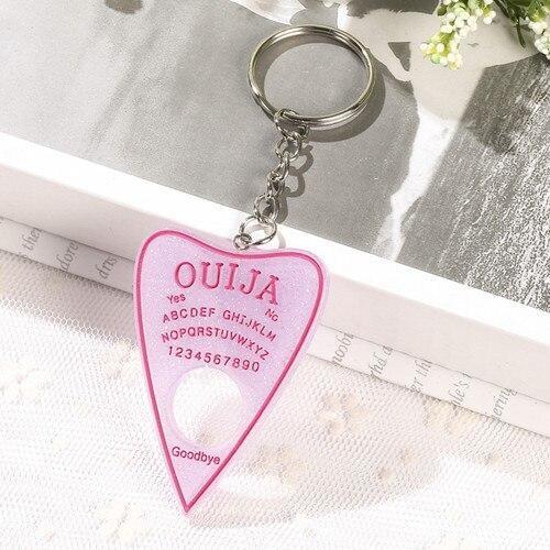 Pale lavender planchette keychain with a bright pink border and lettering, showing the alphabet, numbers and circular cutout beside an open book.