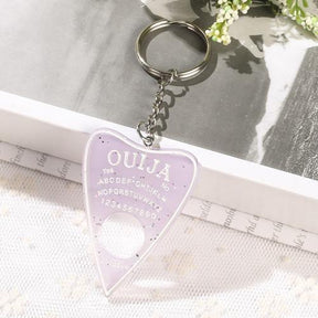 Pale lavender planchette keychain with white lettering, scattered sparkling flecks and a circular opening, displayed on lace beside an open book and small white flowers.