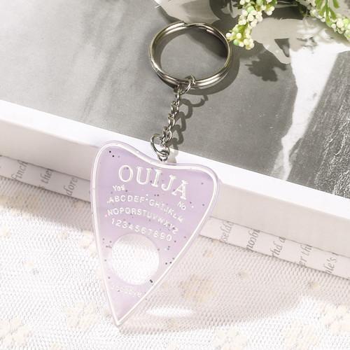 Pale lavender planchette keychain with white lettering, scattered sparkling flecks and a circular opening, displayed on lace beside an open book and small white flowers.