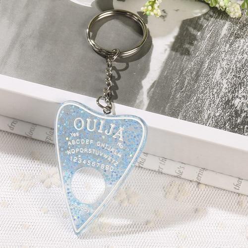 Light blue planchette keychain filled with reflective glitter, with a white outline and lettering. The silver-tone chain drapes over an open book behind it.