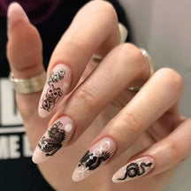 Almond-shaped beige and pale pink press-on nails worn on a hand, with black dragon, lucky-cat and floral line drawings.