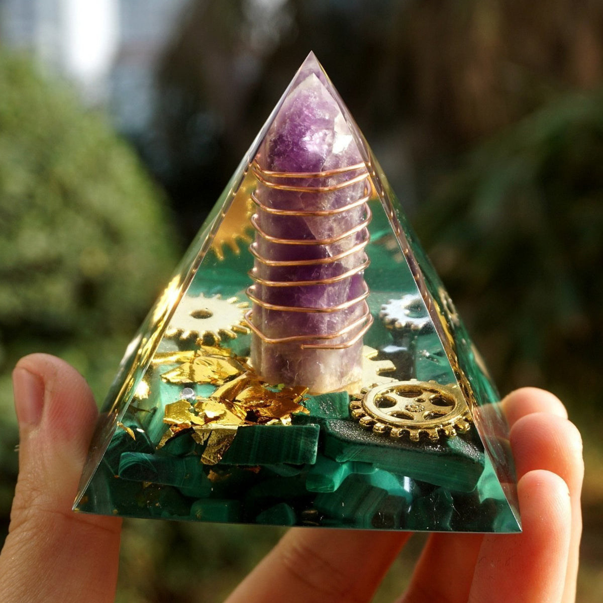 Close front view of a malachite orgone pyramid with an encased purple amethyst point, spiral metal wrapping and gear details.