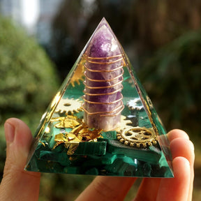 Close front view of a malachite orgone pyramid with an encased purple amethyst point, spiral metal wrapping and gear details.