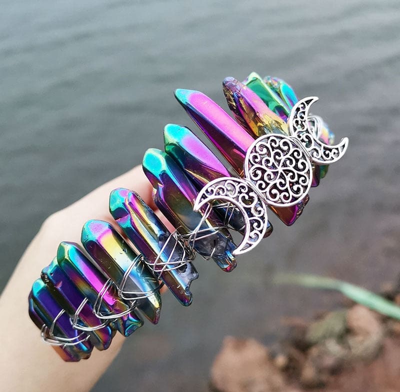 Angled view of a metallic rainbow crystal point crown with a silver-tone triple moon emblem and wire-wrapped band.