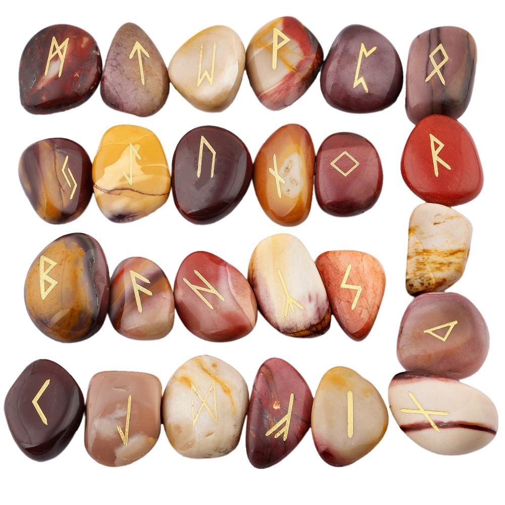 Mookaite rune stone set laid out in rows, showing individual gold-coloured characters and one unmarked stone against mustard, red and beige surfaces.