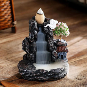 Full view of the mountain-village incense burner on a wooden table, showing the dark rocky outline, terracotta-colored roof, pink blossoms and cascading smoke.
