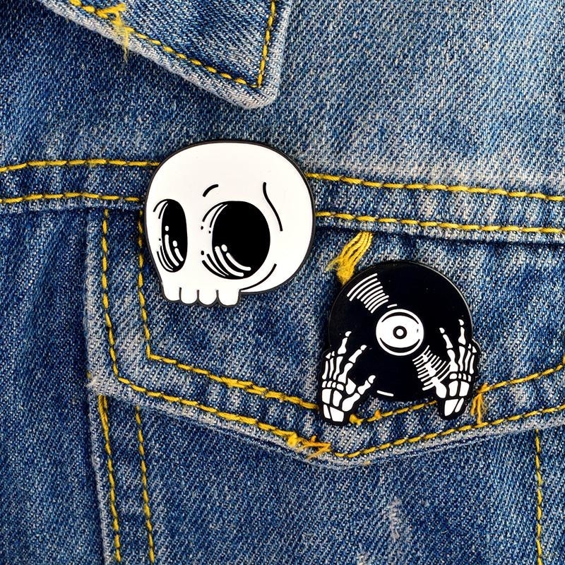 Skull and record-hands enamel pins attached beside the pocket and seams of a blue denim jacket with yellow stitching.
