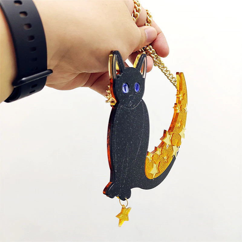Angled close view of the black acrylic cat pendant held by its gold-tone chain, showing the layered edge of the glitter moon and the separate star charm hanging below.