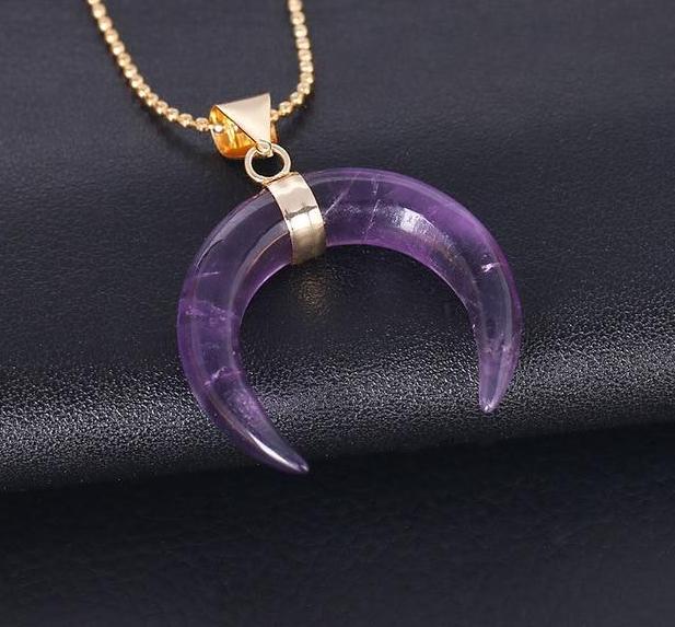 Purple amethyst crescent pendant on a dark background, with a gold-tone band at its centre and a fine ball chain.