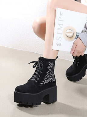 Black platform ankle boots with white pentagram, moon and rose embroidery shown in a cropped crouching outfit view.