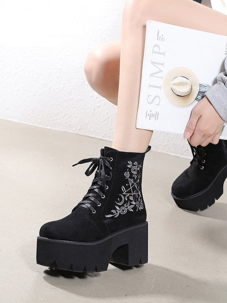 Black platform ankle boots with white pentagram, moon and rose embroidery shown in a cropped crouching outfit view.