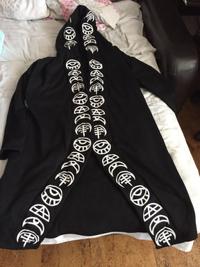 Open-front black cardigan laid flat on bedding, with rows of white symbols running from the hood down both draped front edges.