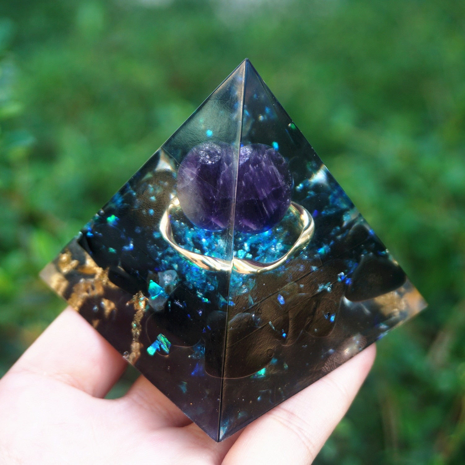Ocean amethyst orgone pyramid with a purple sphere, blue flakes and dark obsidian stones. Angled view on a palm showing the depth of the blue and black layers.