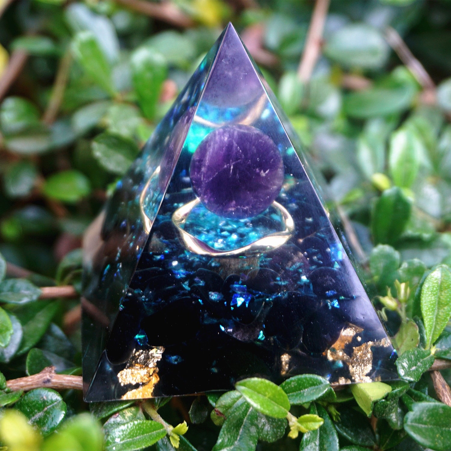 Ocean amethyst orgone pyramid with a purple sphere, blue flakes and dark obsidian stones. Standing among green leaves with the purple sphere and blue layers facing forward.