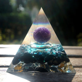 Additional view of Ocean amethyst orgone pyramid with a purple sphere, blue flakes and dark obsidian stones. A second sunlit display view shows the pyramid resting on wood.