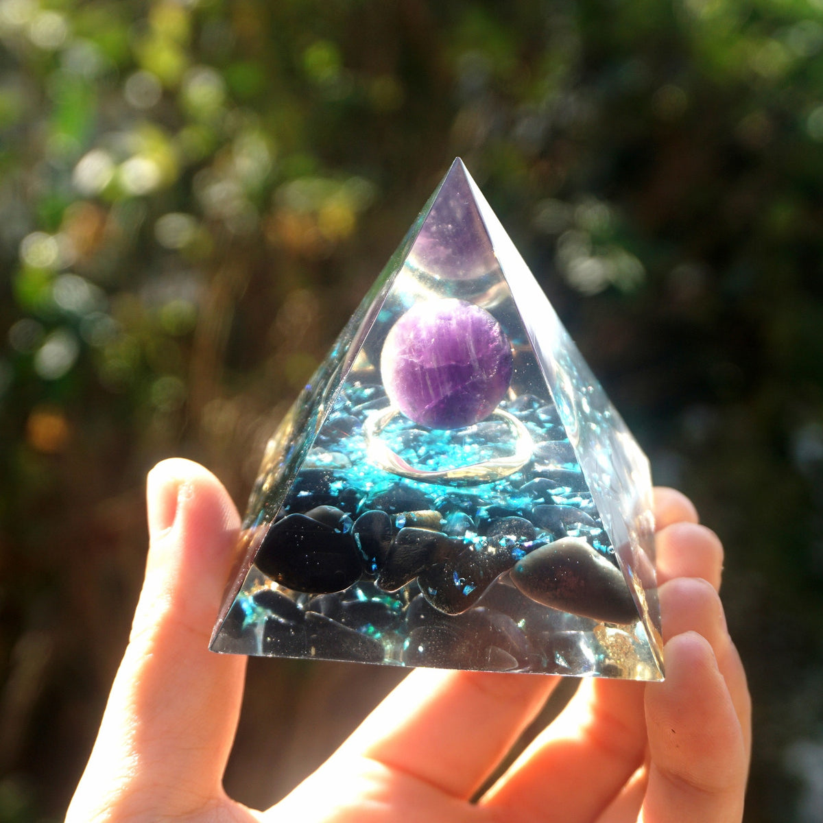 Ocean amethyst orgone pyramid with a purple sphere, blue flakes and dark obsidian stones. Front view in sunlight showing a circular copper detail beneath the sphere.