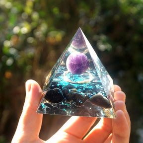 Ocean amethyst orgone pyramid with a purple sphere, blue flakes and dark obsidian stones. Front view in sunlight showing a circular copper detail beneath the sphere.