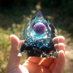 Ocean amethyst orgone pyramid with a purple sphere, blue flakes and dark obsidian stones. Held at arm’s length with the full pyramid and layered stone arrangement visible.