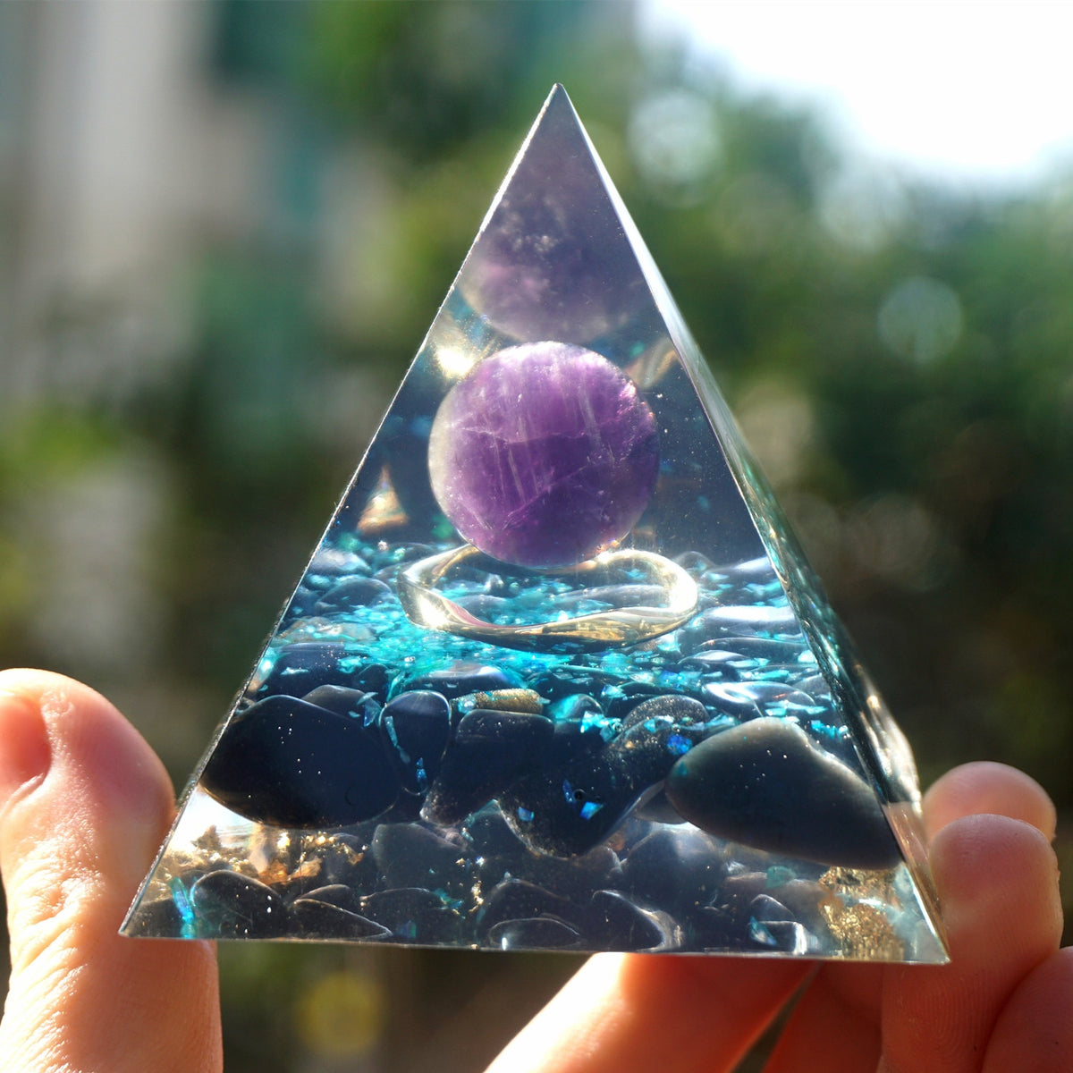 Ocean amethyst orgone pyramid with a purple sphere, blue flakes and dark obsidian stones. Held upright outdoors with the sphere and dark base clearly visible.