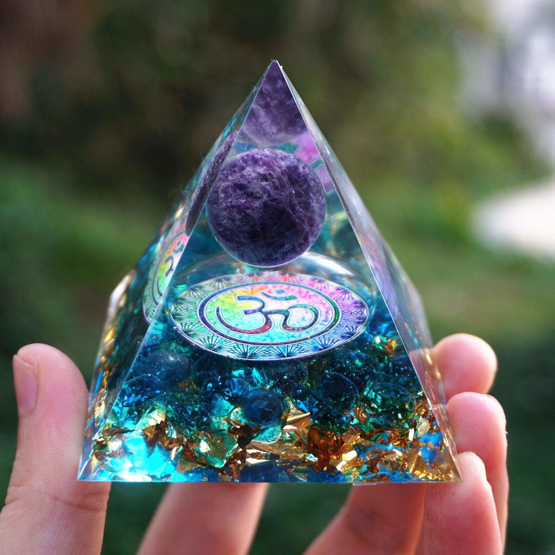 Front view of the amethyst Om pyramid held close, with the metallic symbol centred over a blue quartz base.