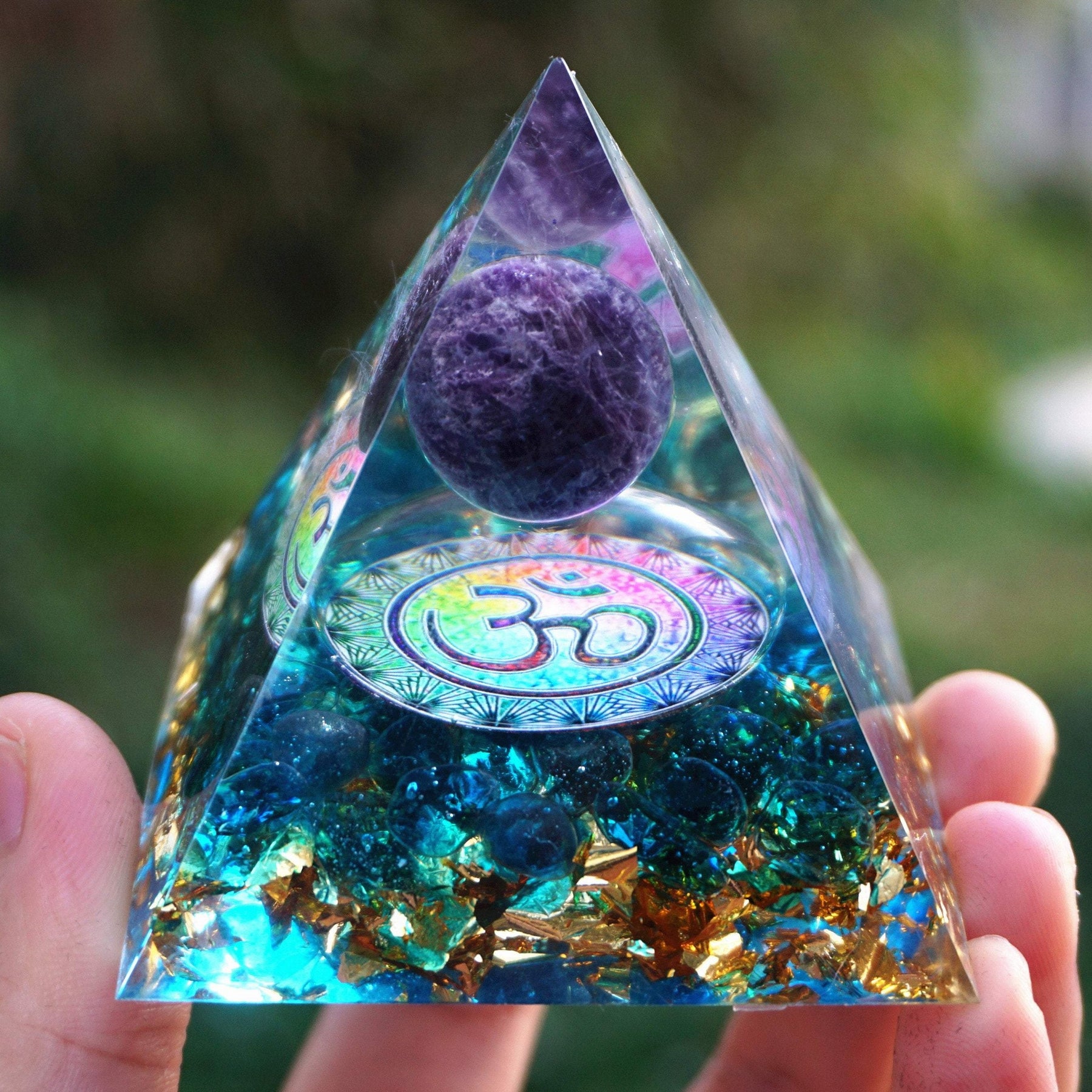 Close view of an Om orgone pyramid with a purple amethyst sphere above a warm metallic symbol and blue quartz base.