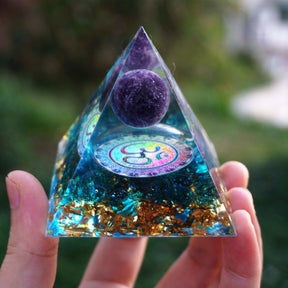An open hand supports the Om pyramid, showing its pointed top and contrasting purple, blue and metallic layers.