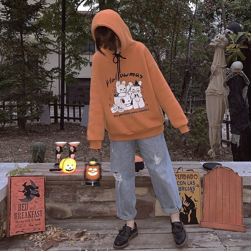 Full outdoor outfit with the orange Hallowmas hood raised, gray trousers and black footwear, photographed among Halloween figures and pumpkins.