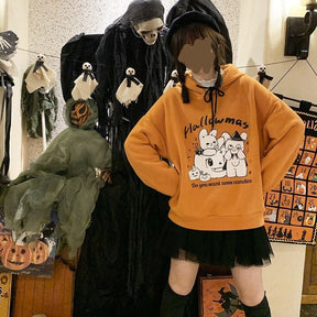 Model wearing the orange Hallowmas hoodie with hands at the waist, showing the broad sleeves and ribbed hem over a black skirt.
