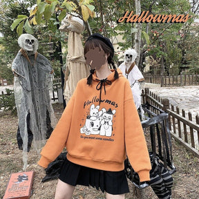 Orange Hallowmas hoodie worn outdoors with a black skirt beside skeleton decorations, with Hallowmas lettering overlaid near the top of the photograph.