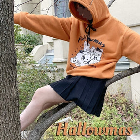 Model seated on a branch wearing the orange graphic hoodie with the hood raised and a black skirt, with Hallowmas text over the outdoor scene.