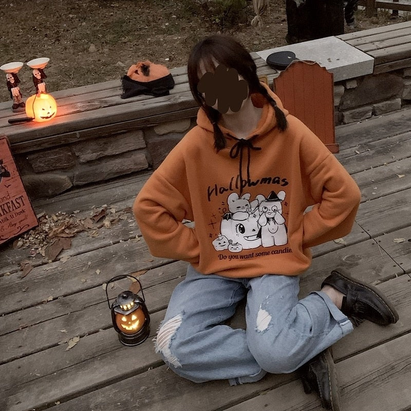 Seated outdoor view of the orange Hallowmas hoodie with gray trousers and a pumpkin lantern on a wooden platform beside the model.