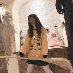Model wearing the orange Halloween graphic sweatshirt and black skirt while holding a broom horizontally in a decorated room.